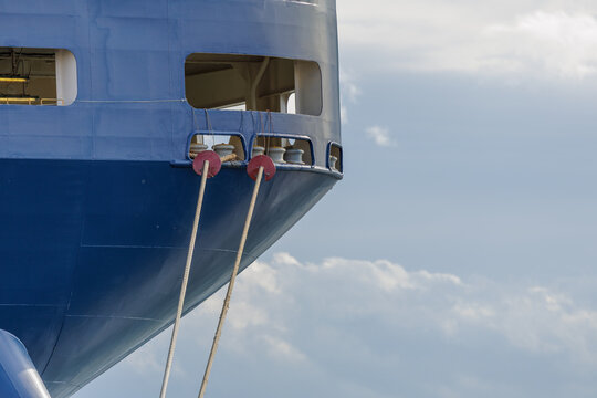 Close-up of a RORO ship's stern with heavy mooring lines running through a chock opening with roller fairleads and secured with red rat guards, set against a cloudy blue sky.