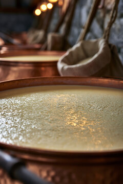 An eye-level, detailed view of fresh milk or whey curdling in large, traditional copper vats during the cheesemaking process, with cheesecloths hanging nearby.