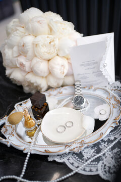 Wedding rings, white peony bouquet, invitation, watch and perfume create an elegant bridal flat lay composition.
