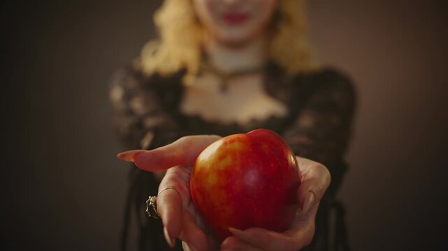 blonde woman dark queen fairytale witch character is giving offering to try poisoned apple. close-up hands gothic fantasy evil girl unrecognizable face holding enchanted red apple for beauty princess