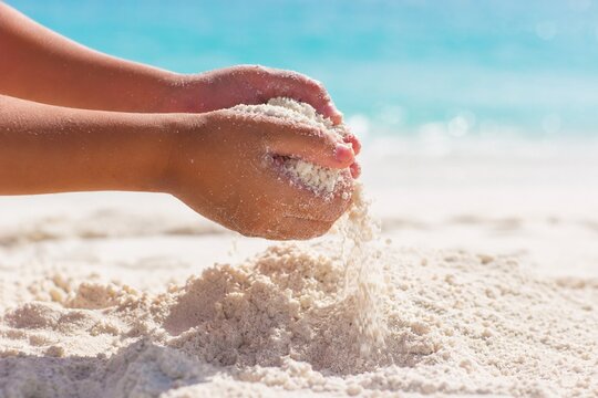 Bright and airy close-up photo of a child's hands letting fine white sand slip through fingers on a sunlit tropical beach with turquoise water, representing sensory play and childhood memories.