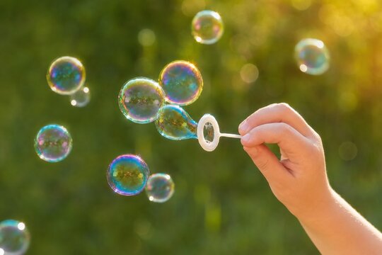 Golden hour lifestyle photography of a child's hand blowing iridescent soap bubbles in a sunlit green park, evoking joyful childhood nostalgia and carefree summer fun.