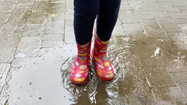 Child in colorful pink rubber boots jumping in a puddle on a rainy day