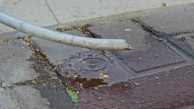 Water dripping from a damaged drainage pipe onto cracked asphalt forming a small puddle on a hot summer day in an urban setting