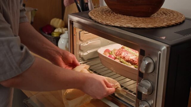 Close-up view of unknown man putting baking dish with meat roast and vegetables into oven, closing door and turning dial to start cooking. Ideal for home appliance marketing or culinary tutorials