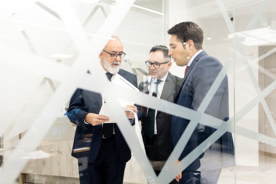 Photograph taken through a glass partition showing a group of three lawyers or businesspeople, aged between 40 and 60, talking to each other using papers in the hallway of a modern office building.