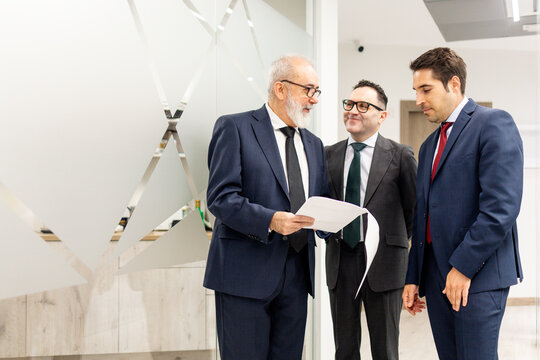 A group of three lawyers or businesspeople, aged between 40 and 60, talking using papers in the hallway of a modern office building. Concept of groups of businesspeople or lawyers.