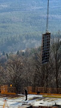 A construction crane lifting heavy concrete formwork panels on a building site overlooking a dense forest, highlighting professional labor and modern development work. Vertical