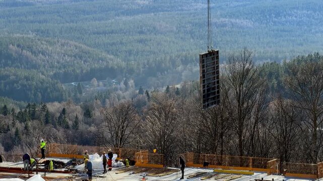A construction crane lifting heavy concrete formwork at a high mountain site, illustrating industrial infrastructure development projects amidst a vast forest landscape scenery. Zoom