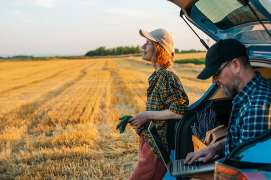 Caucasian farmer couple managing agribusiness online at sunset. Husband working on laptop while wife in cap looking at field.