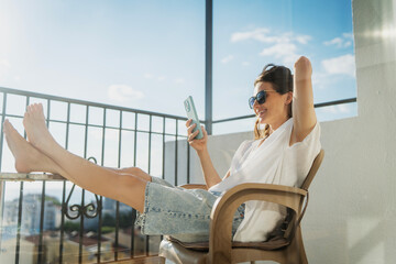 Obraz premium Happy young woman sitting on balcony chair with barefoot legs on table and looking at smartphone screen.