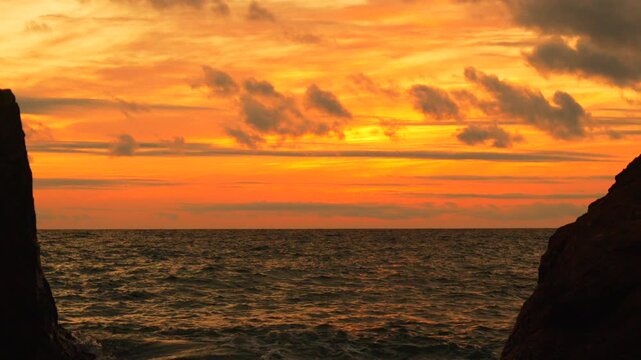 Sunset, ocean, seascape, dramatic orange and gold clouds illuminate rippling dark water framed by coastal rocks.