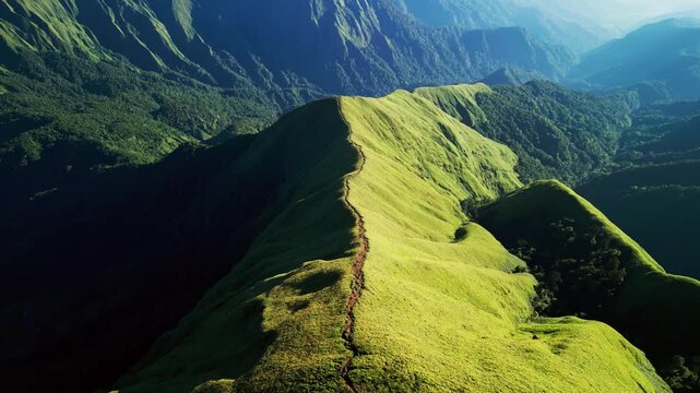 Dramatic light and shadow define a curving grassy mountain spine as a narrow trail winds along the ridge above deep tropical valleys aerial drone footage
