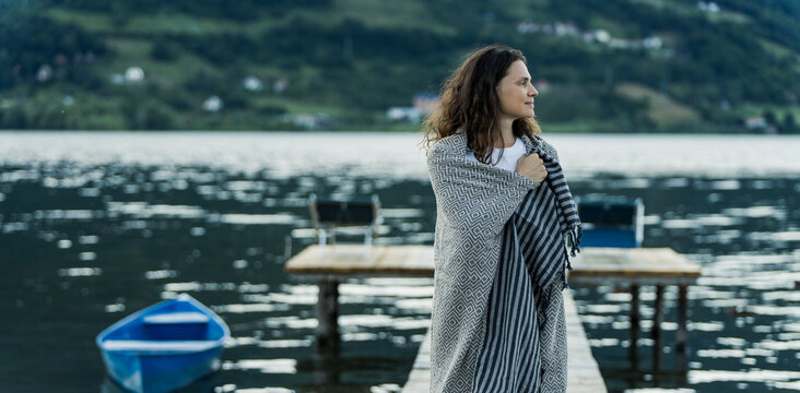Beautiful young woman wrapped in warm blanket standing on wooden pier by scenic lake.