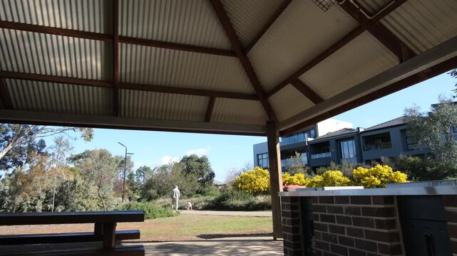 A park pavilion with a corrugated iron roof and BBQ, overlooking a peaceful public parkland in Melton, Melbourne Australia. &nbsp;Outside, a person walks a dog along a footpath near some suburban houses.