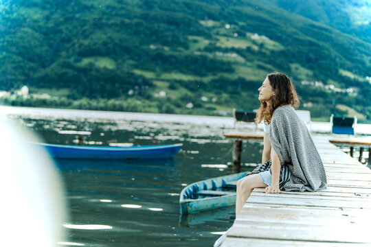 Side view of peaceful young woman wrapped in blanket sitting on wooden pier and looking at mountain lake.