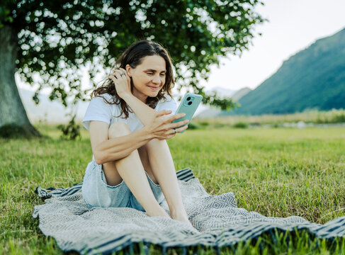Focused female traveler using mobile phone while relaxing in countryside park during summer vacation.
