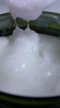 Close up of thick white cream in a container being stirred with a transparent spoon. Rich creamy texture, skincare or cosmetic product mixing concept.