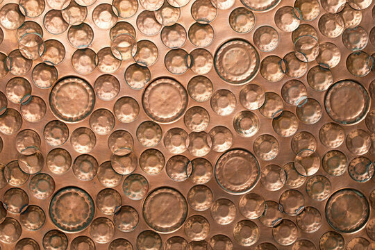Metallic copper surface with a hammered texture featuring various sizes of circular indentations creating a reflective industrial pattern