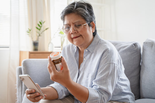 Anxious Asian senior woman holding medicine bottle and smartphone on sofa at home, checking drug information online, reading prescription or using telehealth application
