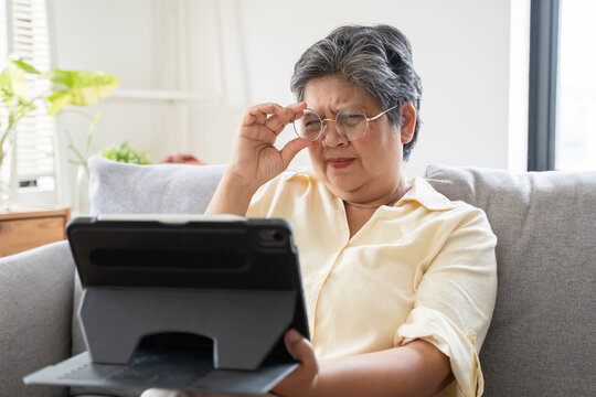 Mature Asian senior woman squinting eyes while looking closely at digital tablet screen on sofa at home, suffering from poor eyesight, presbyopia, vision problems or eye strain concept