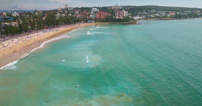 Crowds of people enjoying sandy beach and turquoise waters with rolling waves, surrounded by the coastal city line buildings under a sunny summer sky, perfect for relaxation and holidays. Aerial view