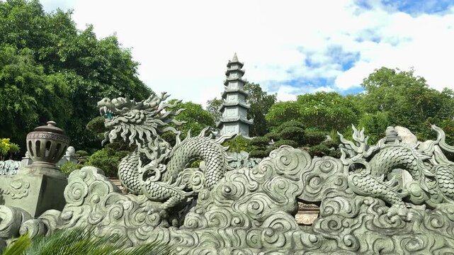 Stone dragon sculptures and a traditional pagoda tower rise against a blue sky, showcasing the detailed artistry and spiritual significance of buddhist architecture at a vietnamese temple