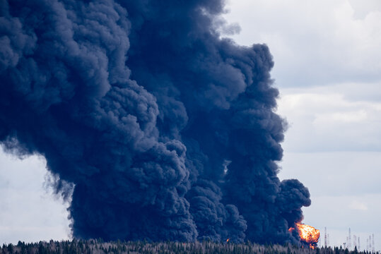 Towering plume of dense black smoke rising from a large oil tank engulfed in flames, indicating a significant industrial accident and environmental pollution