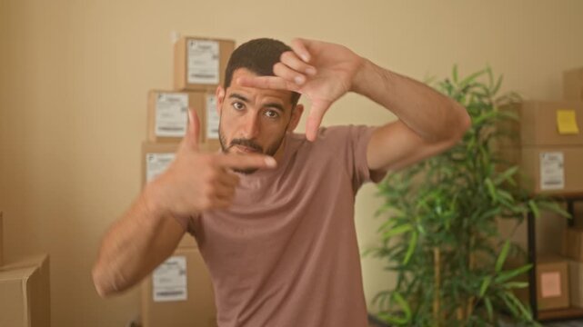 Hispanic man frames camera with hands and points at cardboard parcels stacked on shelves in a small business building; confidence.