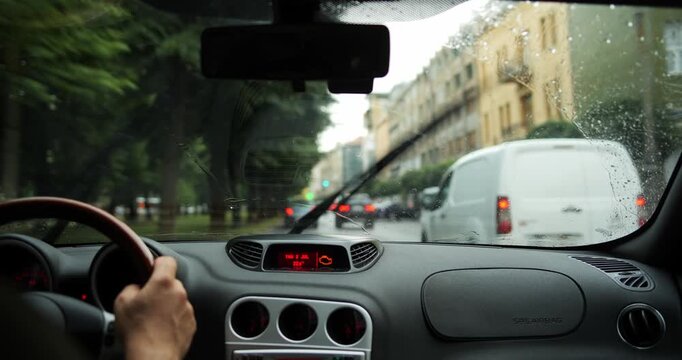 Inside car driving on rainy city street past buildings and trees with raindrops on windshield and dashboard in shallow depth of field focus