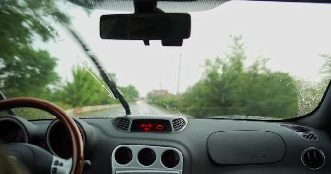 Car interior POV driving on rainy street past residential houses and village buildings with windshield wipers wiping raindrops