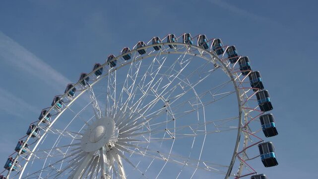 Low-angle view of a large white observation wheel with enclosed passenger cabins and geometric steel framework against a clear blue sky