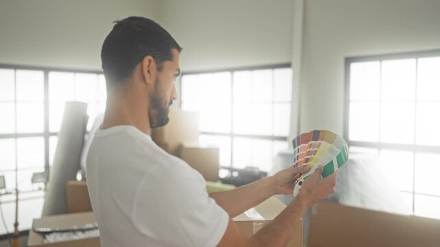 Man holding a cardboard box and flipping paint swatches while examining a color palette in a bright building; planning focus.