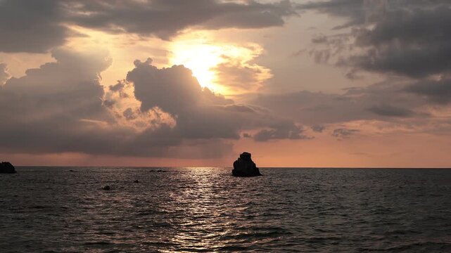 Sun breaking through storm clouds over a silhouetted sea stack at dusk, Tyrrhenian Sea near Tropea, Calabria, Italy