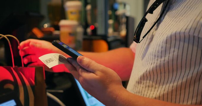 Mobile payment at coffee counter with smartphone and receipt in hand, customer taps to pay while holding shopping bag, close up contactless checkout in cafe, quick, convenient purchase interaction.
