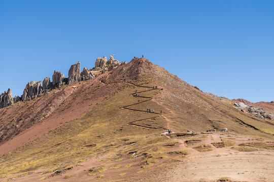 A group of hikers ascends a steep, zigzagging dirt trail leading to the summit ridge of Palccoyo Rainbow Mountain, Peru, with sharp rock formations in the background