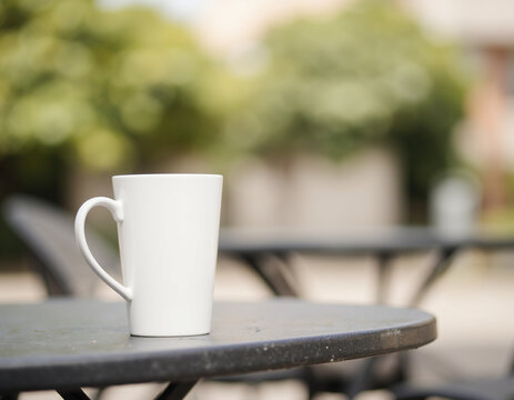 Blank Tall White Ceramic Coffee Mug Mockup Placed on Outdoor Cafe Table with Blurred Background
