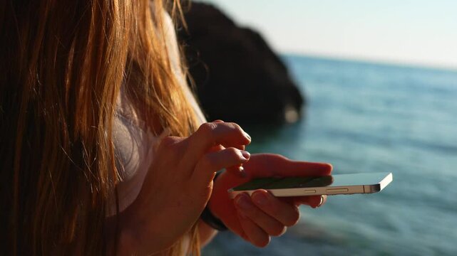 Smartphone beach hands close up of a young woman with long red hair using her mobile device while standing on a coastal shore