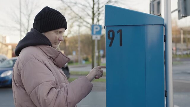 Young woman wearing a winter jacket and knitted beanie presses a button on a blue automated parking terminal beside a roadside parking area.