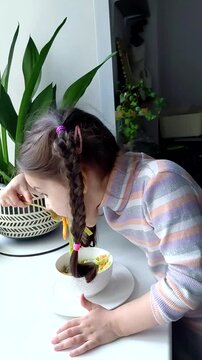 A little girl eats pasta with vegetables near the window. Funny child eats vegetables and laughs