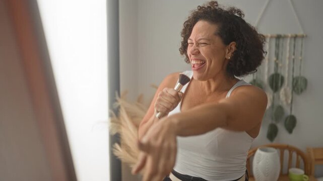 Woman holding makeup brush sings into it and points toward mirror while laughing in studio; joy self care.