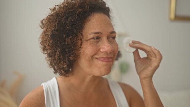Woman smiles and wipes face with cotton pad, dabs cheek and removes makeup in bedroom; gentle self care calm.