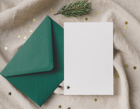 Empty white greeting card and open dark green envelope with golden star confetti and pine needles on a neutral textured textile, perfect for winter holiday mockups