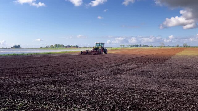 Green tractor plows dark soil, creating straight furrows. Field displays distinct dark and light rows of plowed earth. Tractor moves steadily across vast farmland under clear sky