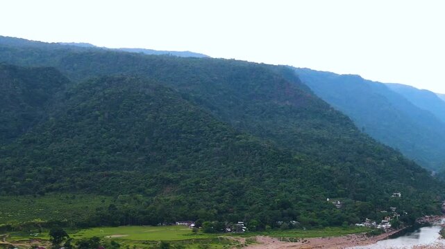Lush Meghalaya hills rise above a dry rocky riverbed and scattered border village houses in a wide daytime drone flyover.