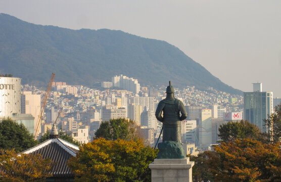 Admiral Yi Sun sin statue standing in Yongdusan Park, providing an elevated view of the sprawling Busan downtown district with numerous buildings and a distant mountain under a clear sky