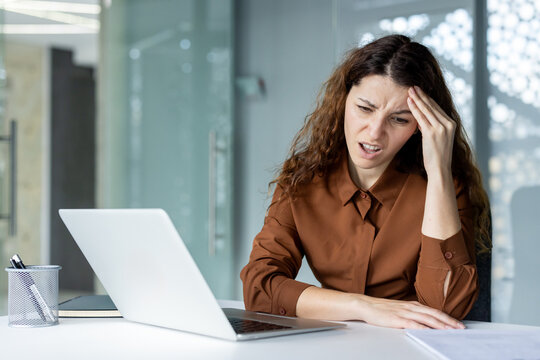 Young businesswoman experiencing a severe headache and high stress while working on her laptop in a modern office, feeling overwhelmed by professional responsibilities and a demanding workload