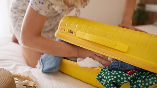 Frustrated young woman trying to forcefully close an overpacked yellow suitcase filled with clothes, pressing down on the luggage while preparing for a summer vacation and holiday trip.