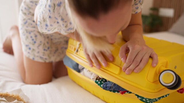 Determined woman struggling to close an overstuffed yellow suitcase full of clothes while kneeling on the bed, getting ready for her much anticipated summer vacation trip
