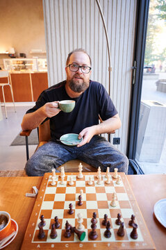 Man facing camera with chessboard and cup, inviting expression and readiness for rematch, casual game setting with glass window view, pastry on plate and warm ambient light, conversational mood